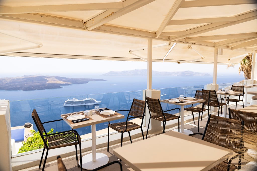 Al fresco dining in Santorini with Oceania Vista® anchored below, where fine cuisine meets unforgettable views.