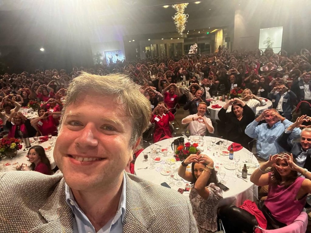 Zak Williams takes a smiling selfie in front of a large seated audience at a formal event, with attendees forming heart shapes with their hands toward the camera.