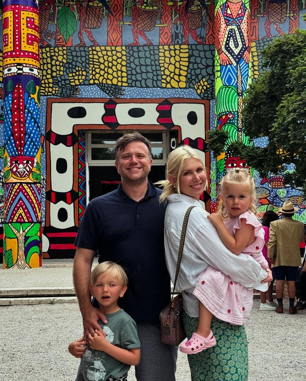 A smiling man and woman pose with their two young children in front of a vibrantly colorful, tribal-patterned building facade. The man holds the hand of a blond boy, while the woman carries a blond girl in a pink dress.