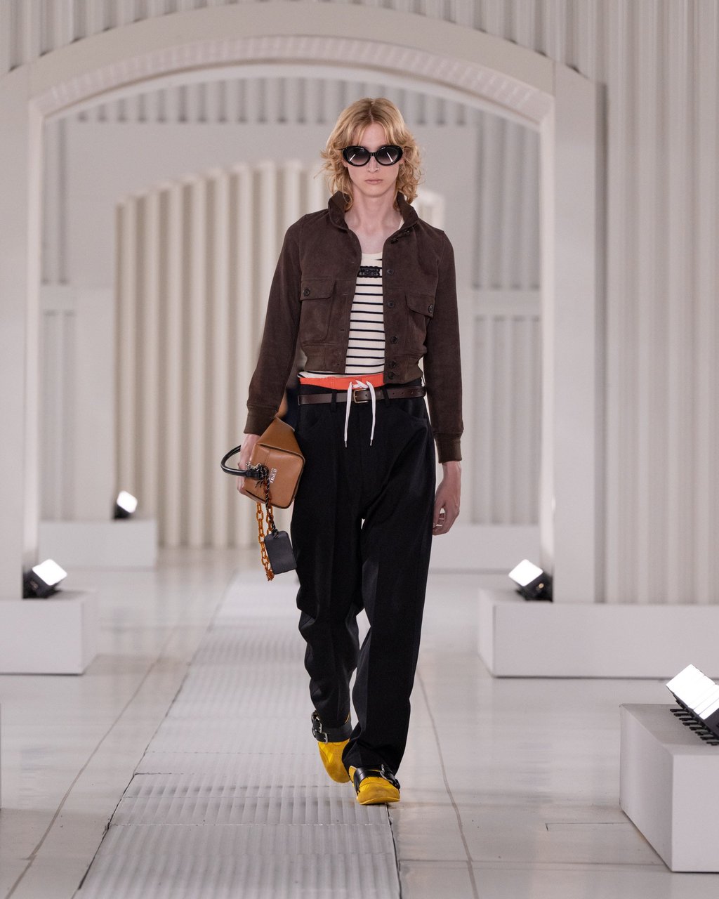 An Onitsuka Tiger spring/summer 2026 look sees a striped top layered under a cropped brown jacket, paired with relaxed black trousers and yellow Mexico sneakers. Photo: Handout
