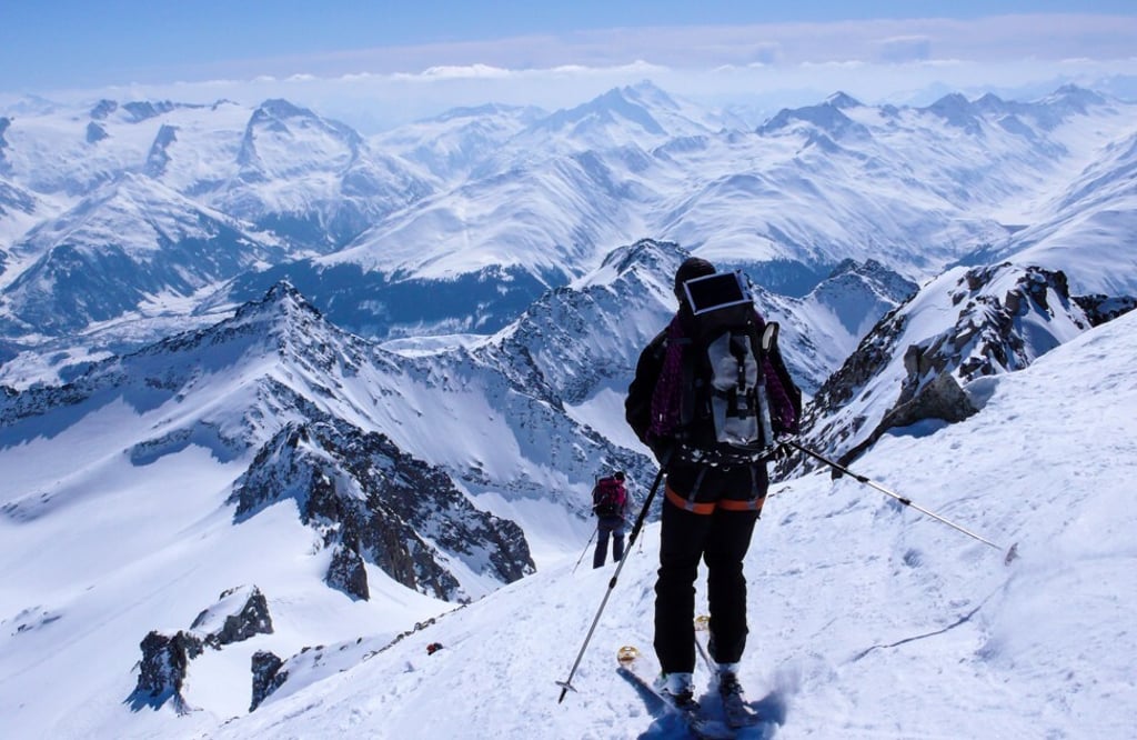 A backcountry skier skiing down from a high alpine summit with a large backpack and solar panel. Photo: Shutterstock.