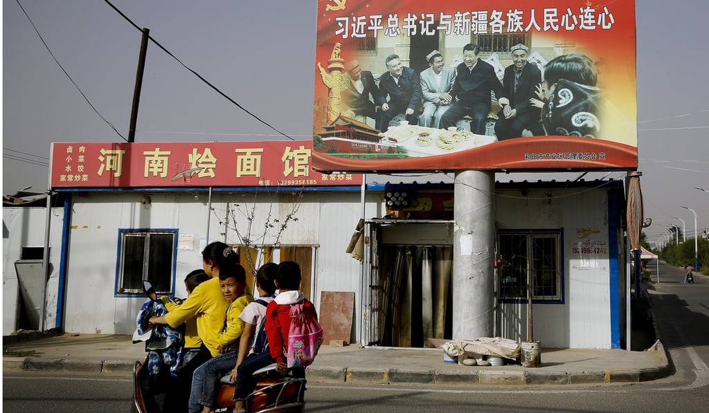 An Uygur woman and school children on a scooter ride past a picture of President Xi Jinping joining hands with a group of Uygur elders at the village. Photo: AP An Uygur woman and school children on a scooter ride past a picture of President Xi Jinping joining hands with a group of Uygur elders at the village. Photo: AP
