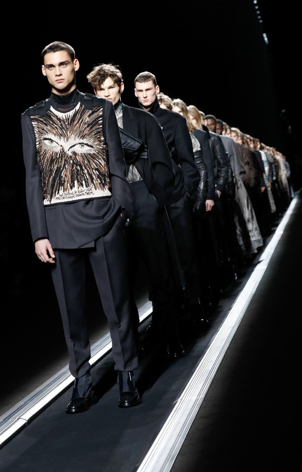 A line of models, including one at the front wearing a utility vest over a dark suit, stand on the moving conveyor-belt catwalk while presenting Dior creations during its men’s fall/winter 2019-20 collection show at Parish Fashion Week. Photo: AFP