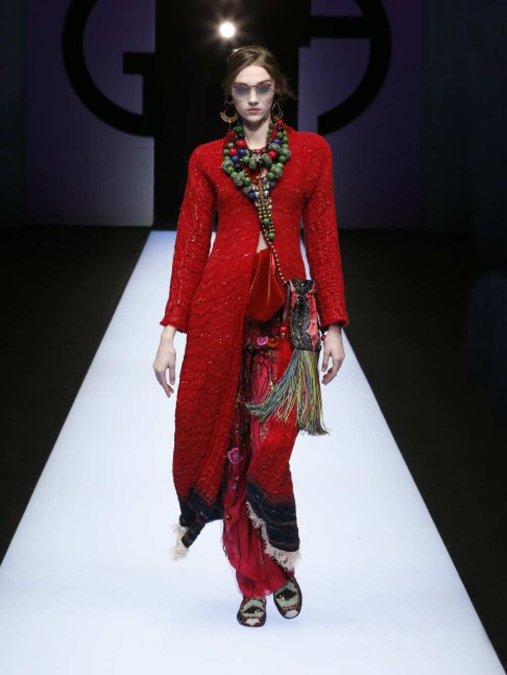 Showstopping long red coat worn with long embroidered sarong-style skirt and multi-coloured fabric moccasins.