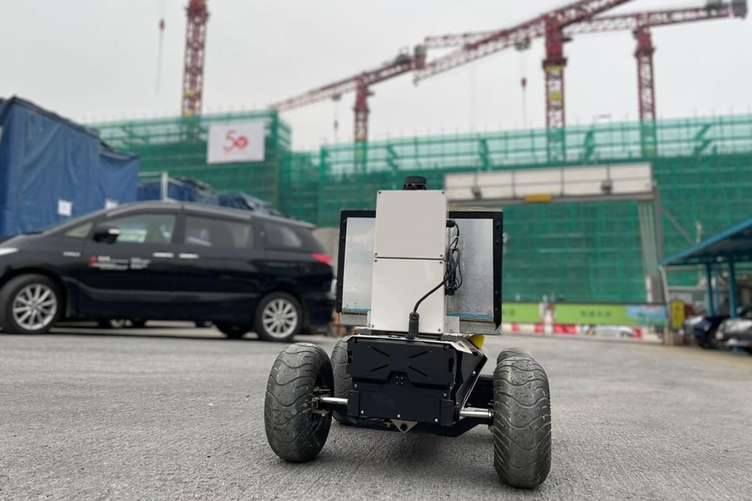 A mobile construction robot with large wheels is positioned on an active construction site in Hong Kong, surrounded by cranes, construction barriers, and ongoing building work.