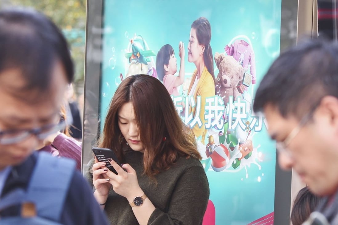 People check their mobile phones in front of a Tmall Singles' Day Global Shopping Festival poster at a bus station in Shanghai on Saturday. Photo: Simon Song