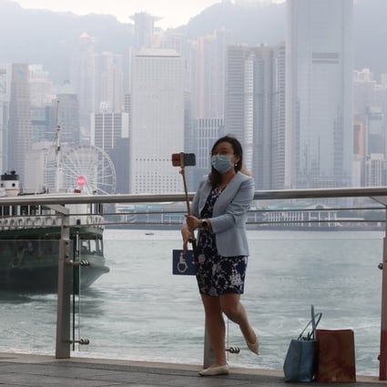 A woman in a face mask with a selfie stick poses for a photo with the Victoria Harbour and Hong Kong skyline in the background. Photo: K.Y. Cheng